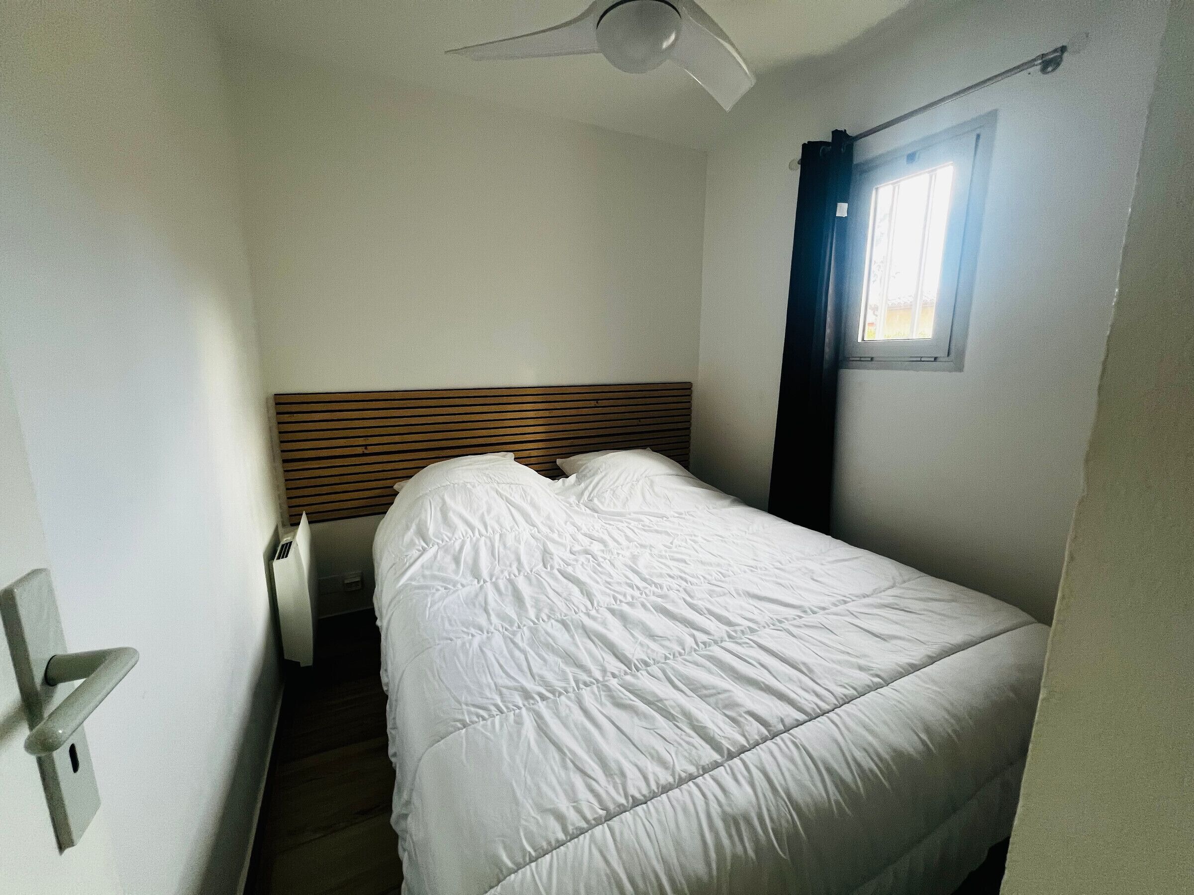 Een kleine kamer met een enkel bed, een houten hoofdeinde, een plafondventilator en een raam met gordijn.
