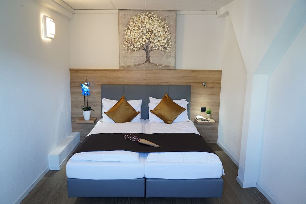 A modern hotel room with a large bed, a wooden headboard, a wall-mounted artwork of a tree, and a bedside table with a lamp and a plant.