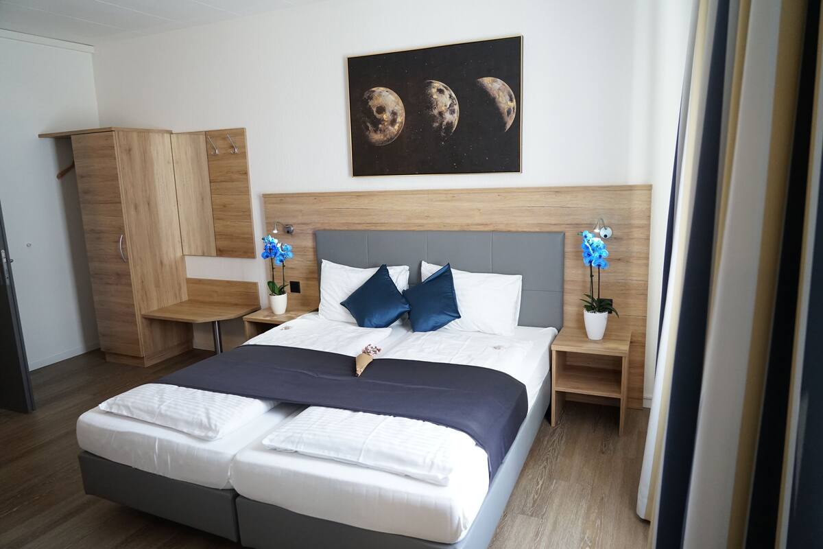 A modern bedroom with a double bed, a wooden wardrobe, a nightstand with a lamp, and a wall-mounted painting of the moon phases.