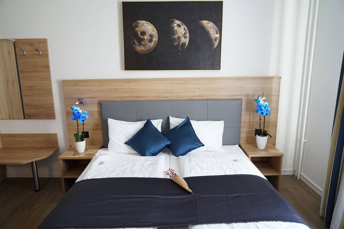 A neatly arranged bedroom with a bed, two nightstands, a wooden headboard, a wall-mounted painting of moons, and two potted plants.