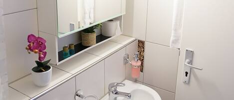 A bathroom with a white sink, mirror, and tiled walls. There is a trash can and a small shelf with toiletries.