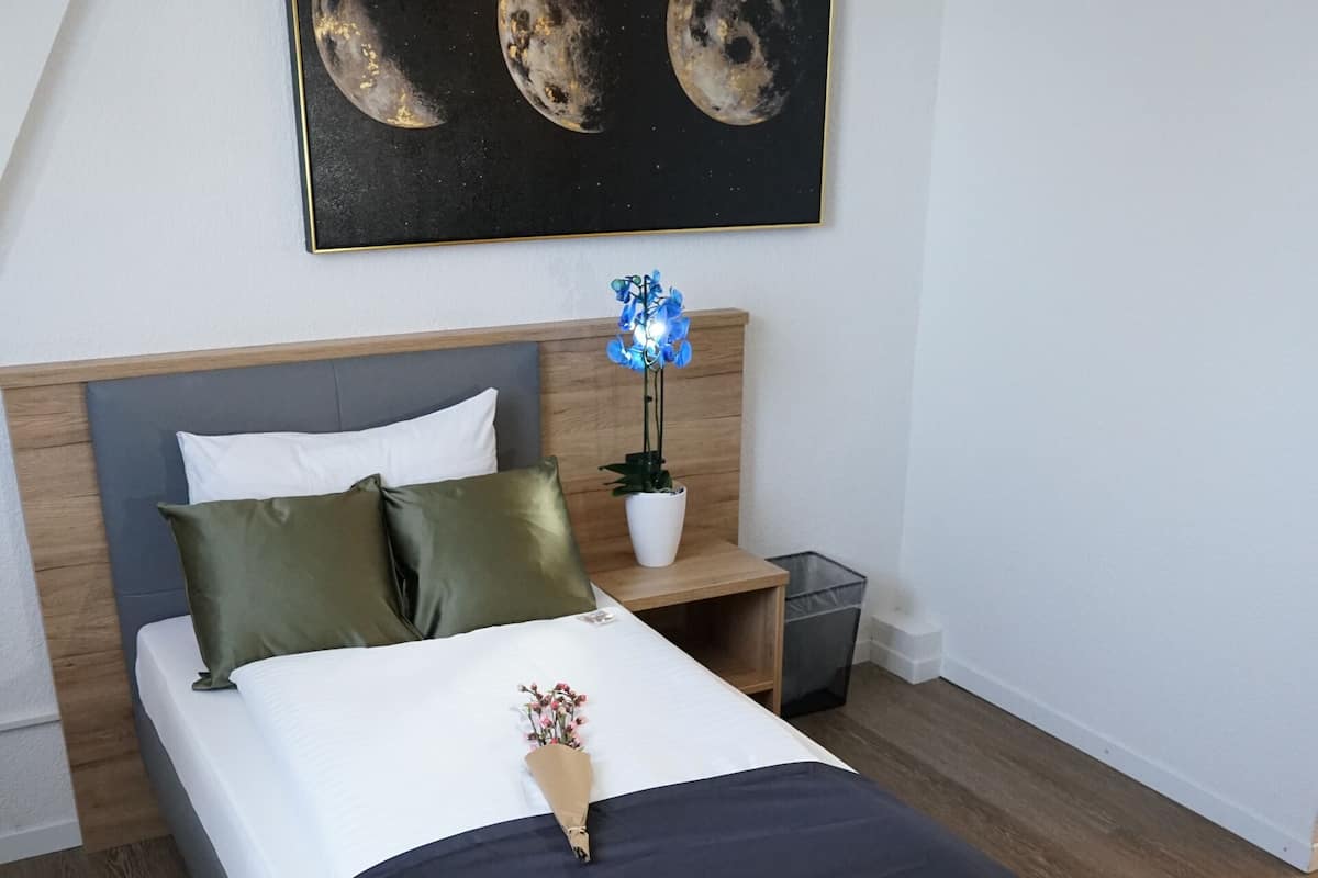 A neatly made bed with a dark blue bedspread and white pillows, a wooden headboard, a nightstand with a potted plant, and a framed picture of the moon phases on the wall.