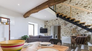 A modern kitchen with a wooden table set for dining, a stone wall, and a staircase with wooden railings. - Charming Stone Barn Near the Sea (Bretteville)