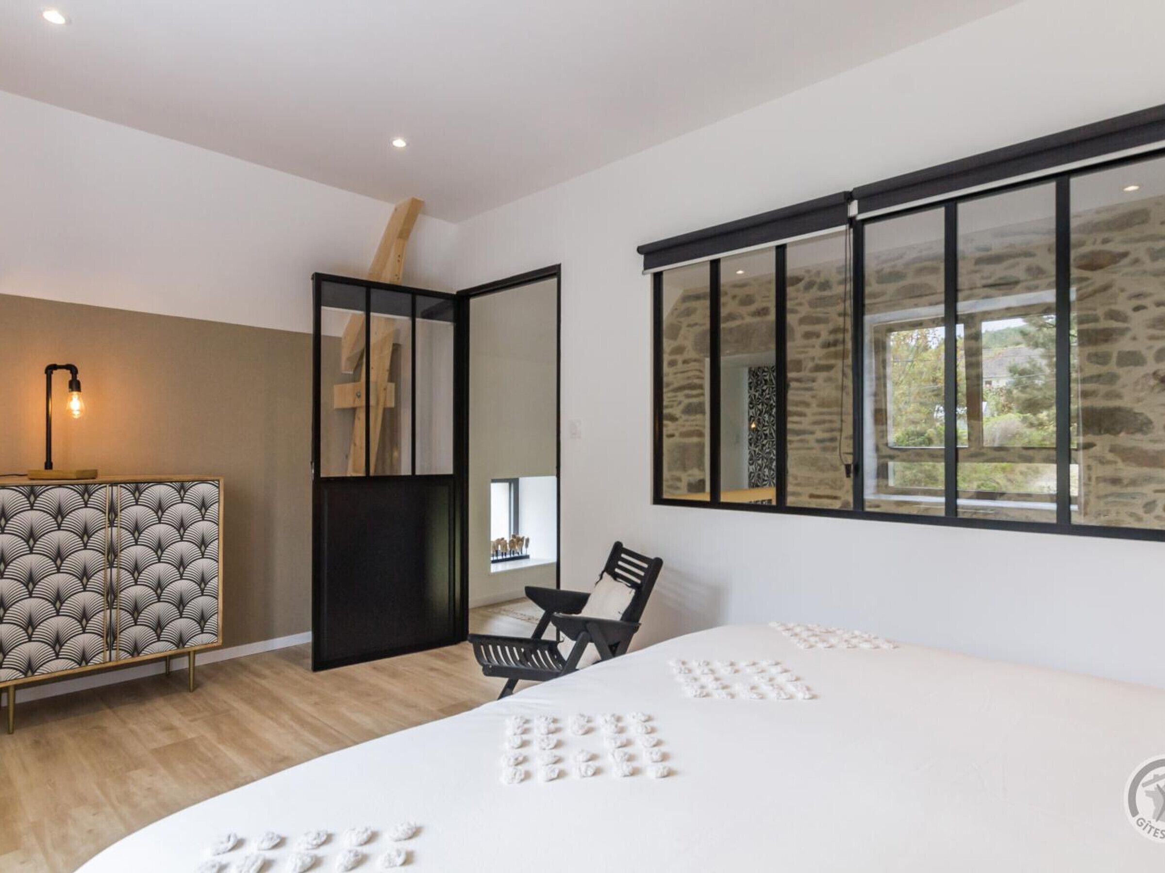 A modern bedroom with a large bed, a desk, a chair, and a window with stone walls.