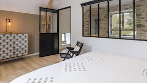 A modern bedroom with a large bed, a desk, a chair, and a window with stone walls. - Charming Stone Barn Near the Sea (Bretteville)