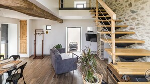 A spacious living area with a wooden staircase, a sofa, a dining table, and a potted plant. - Charming Stone Barn Near the Sea (Bretteville)