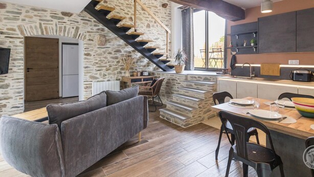 A modern kitchen with a stone wall, wooden staircase, and a dining area with a sofa and chairs. - Charming Stone Barn Near the Sea (Bretteville)
