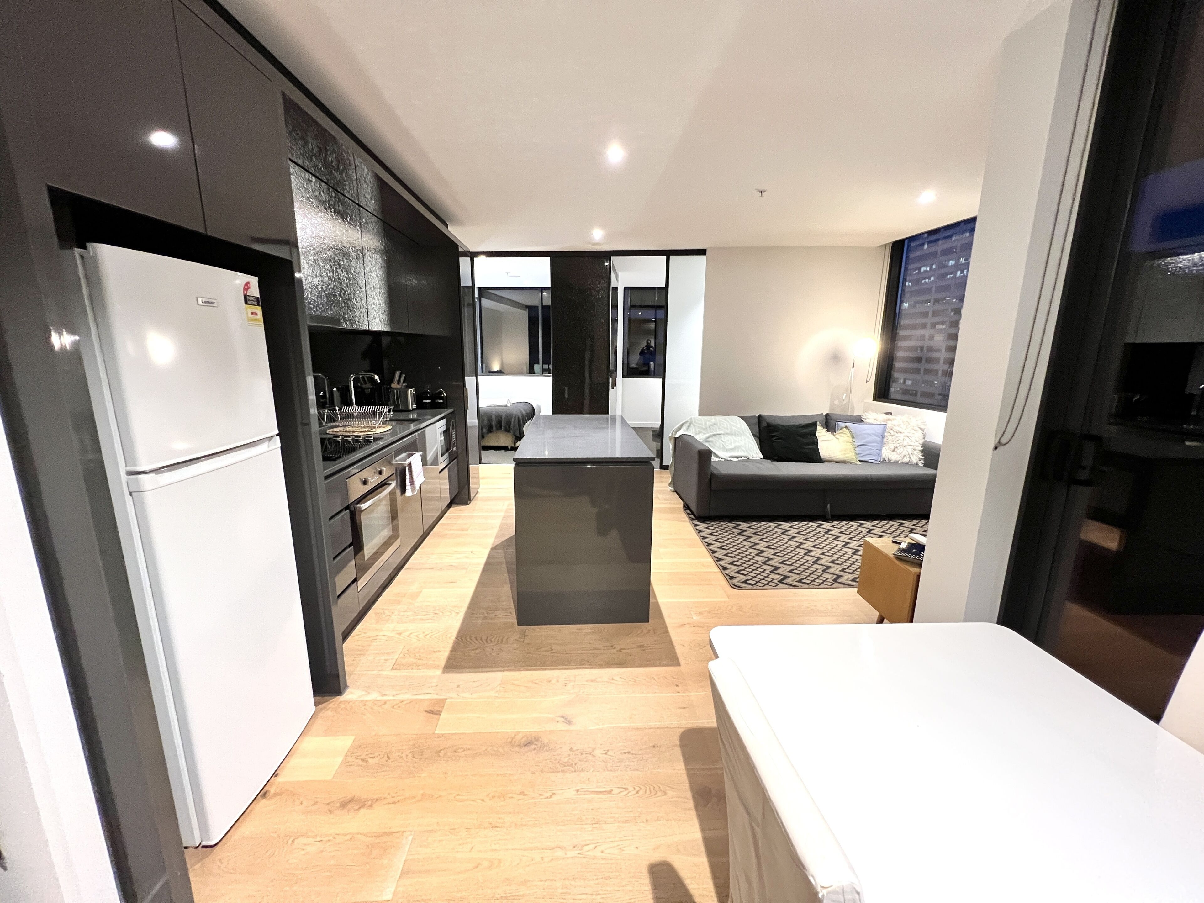 A modern kitchen with a refrigerator, oven, and microwave. There is a dining area with a table and chairs. The room has a large window and a sofa.