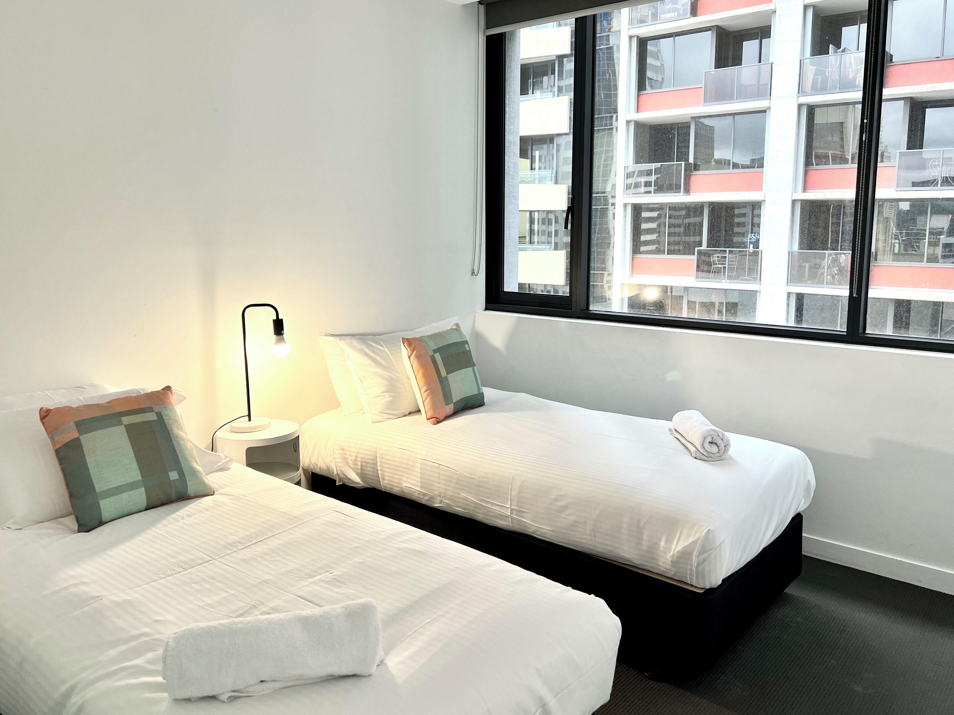 A hotel room with two beds, a window with a view of buildings, and a bedside lamp.
