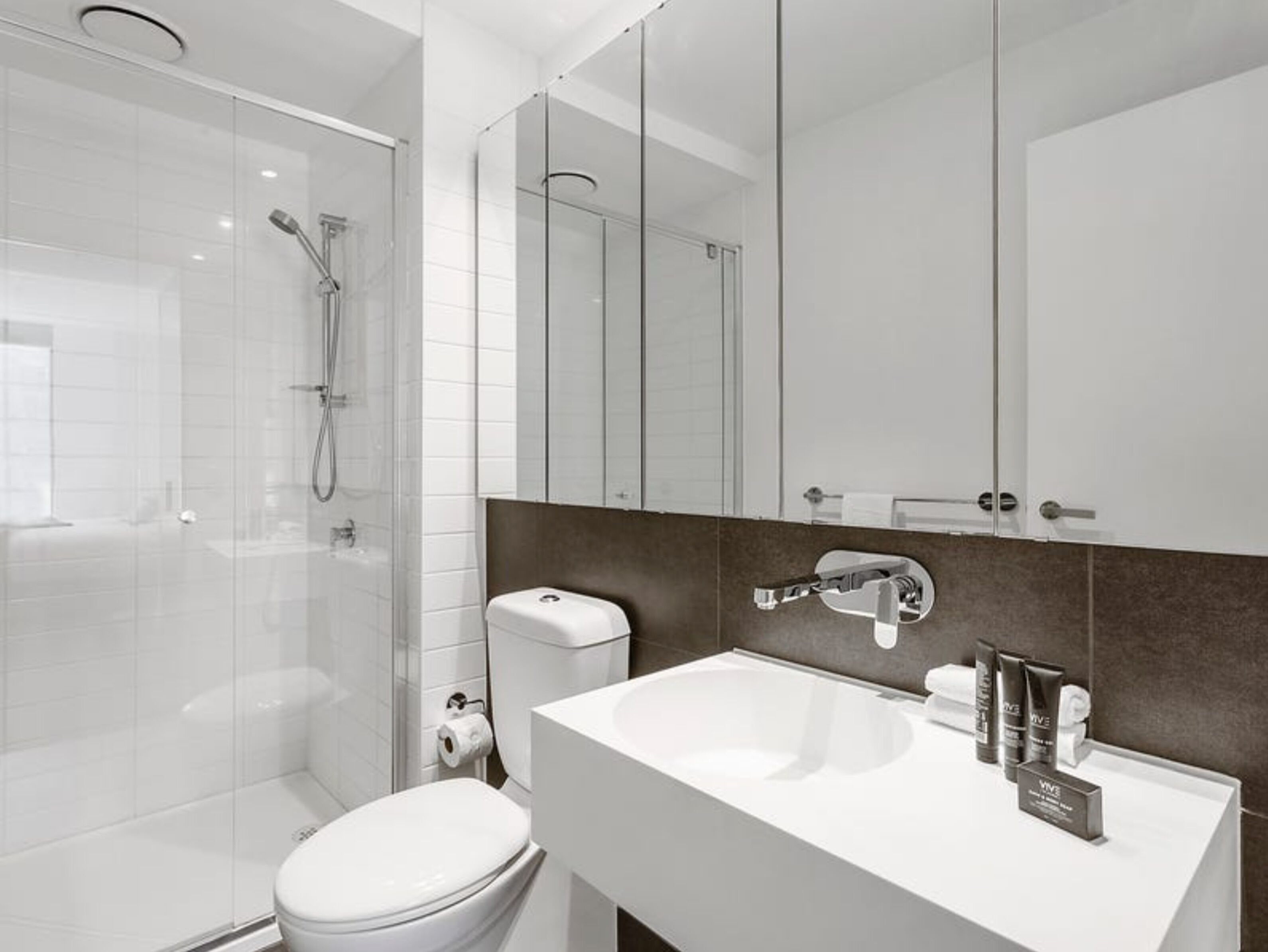 Un baño moderno con cabina de ducha de vidrio, inodoro blanco, tocador con espejo y lavamanos.