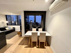 A modern living room with a dining area, a sofa, and a view of a cityscape through large windows. - ReadySet Apartments at Manhattan (Melbourne)