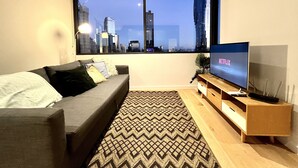 A modern living room with a large window, a grey sofa, a patterned rug, and a wooden TV stand with a flat-screen TV displaying the Netflix logo. - ReadySet Apartments at Manhattan (Melbourne)