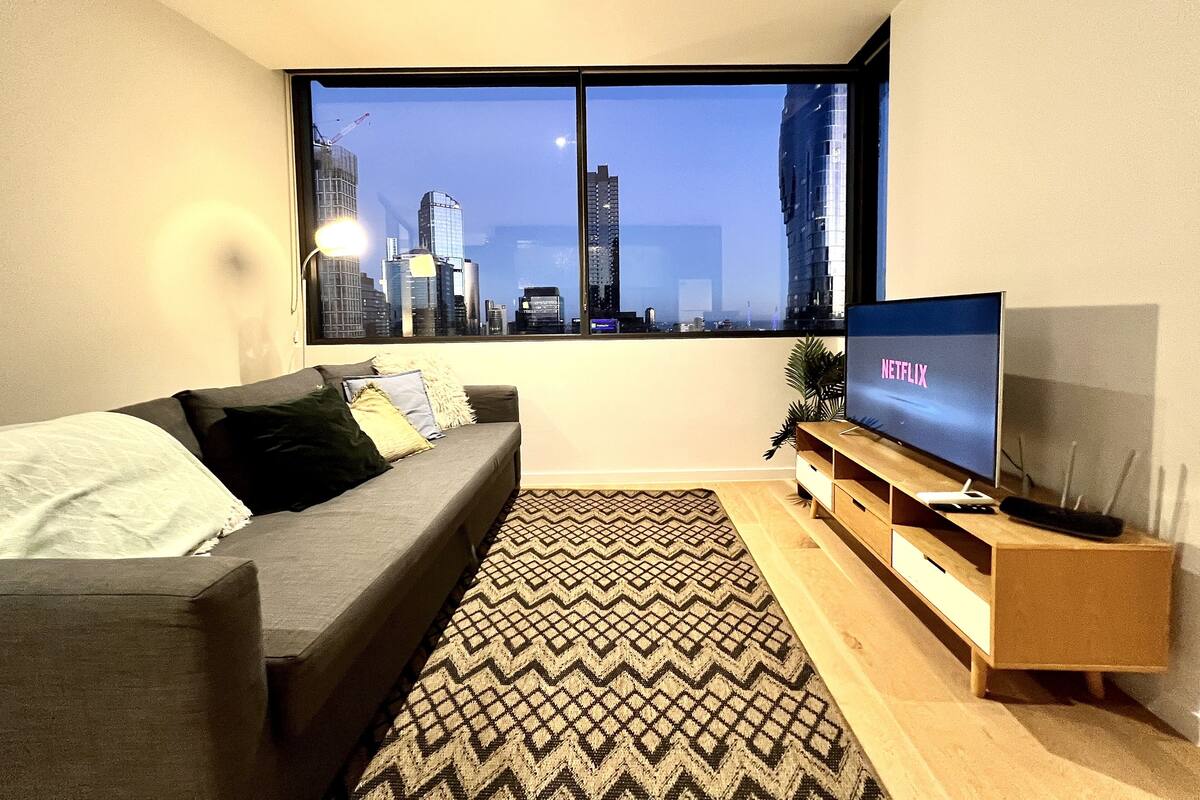 Un salon moderne avec une grande fenêtre, un canapé gris, un tapis à motifs et un meuble TV en bois sur lequel est posée une télévision à écran plat affichant le logo de Netflix.