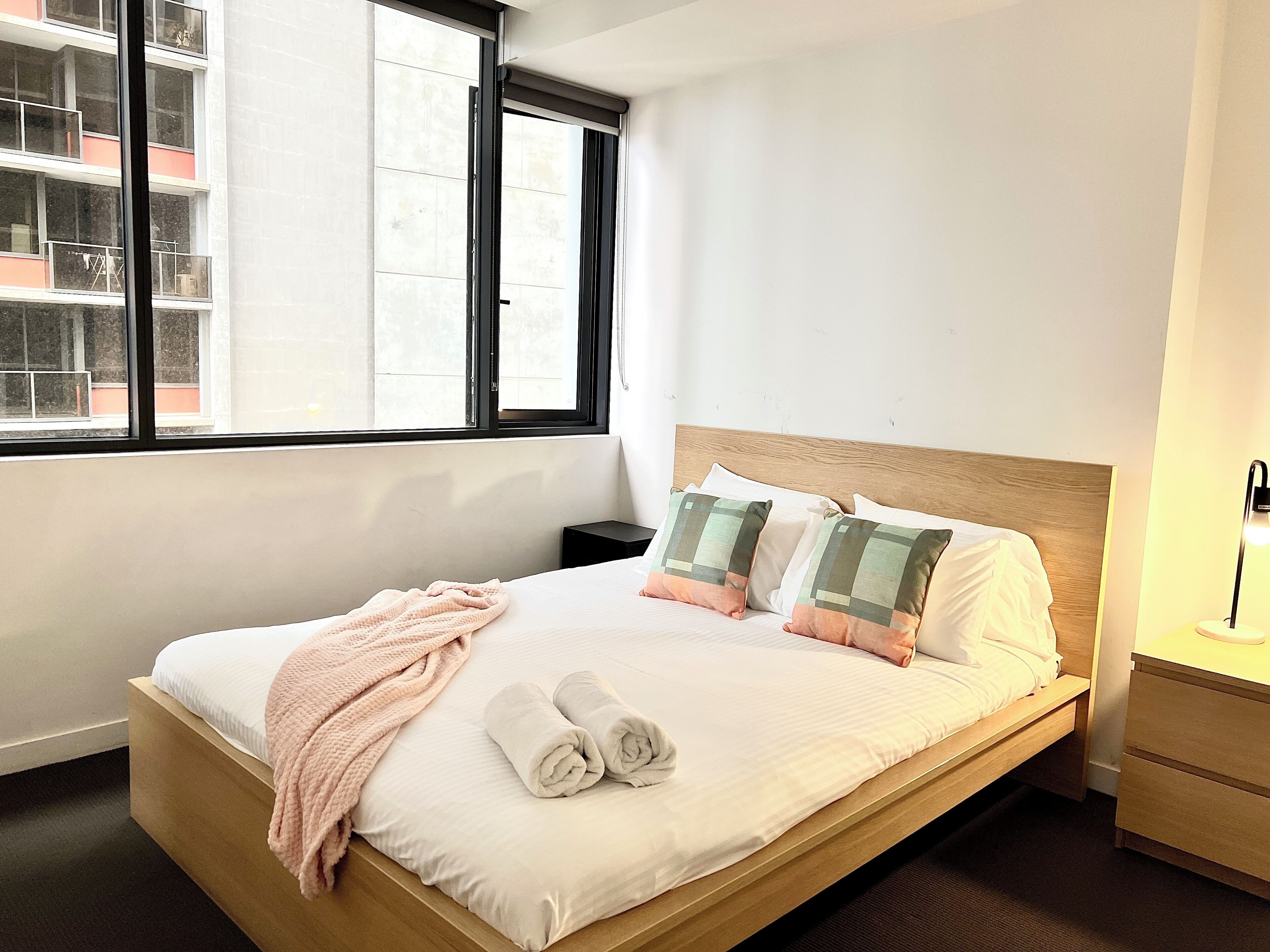 A neatly made bed with a wooden headboard, a pink blanket, and two rolled towels.