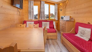 Un interno accogliente di una baita con un tavolo da pranzo in legno, due panche rivestite di rosso e bianco, un piccolo mobiletto in legno e un televisore montato a parete.