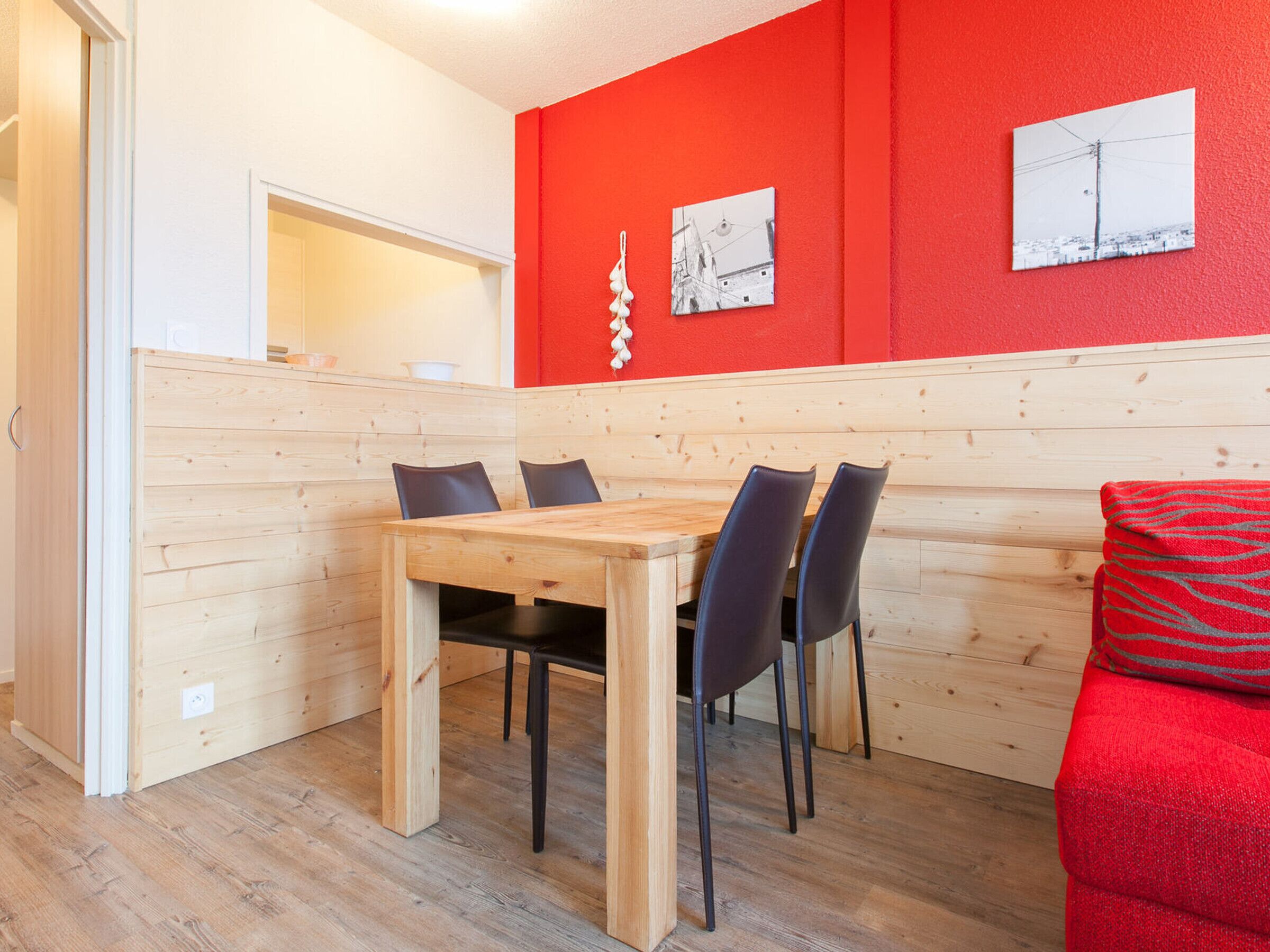 A dining area with a wooden table and chairs, a red wall with two framed pictures, and a red sofa.