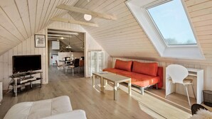 Un loft spacieux avec un plafond haut, des poutres en bois, un canapé orange confortable, un fauteuil blanc et un téléviseur dans l’espace de vie.