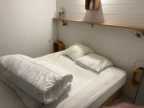 Un lit bien fait, avec une literie blanche, une table de chevet avec une lampe et une étagère au-dessus du lit, ornée d’objets décoratifs.