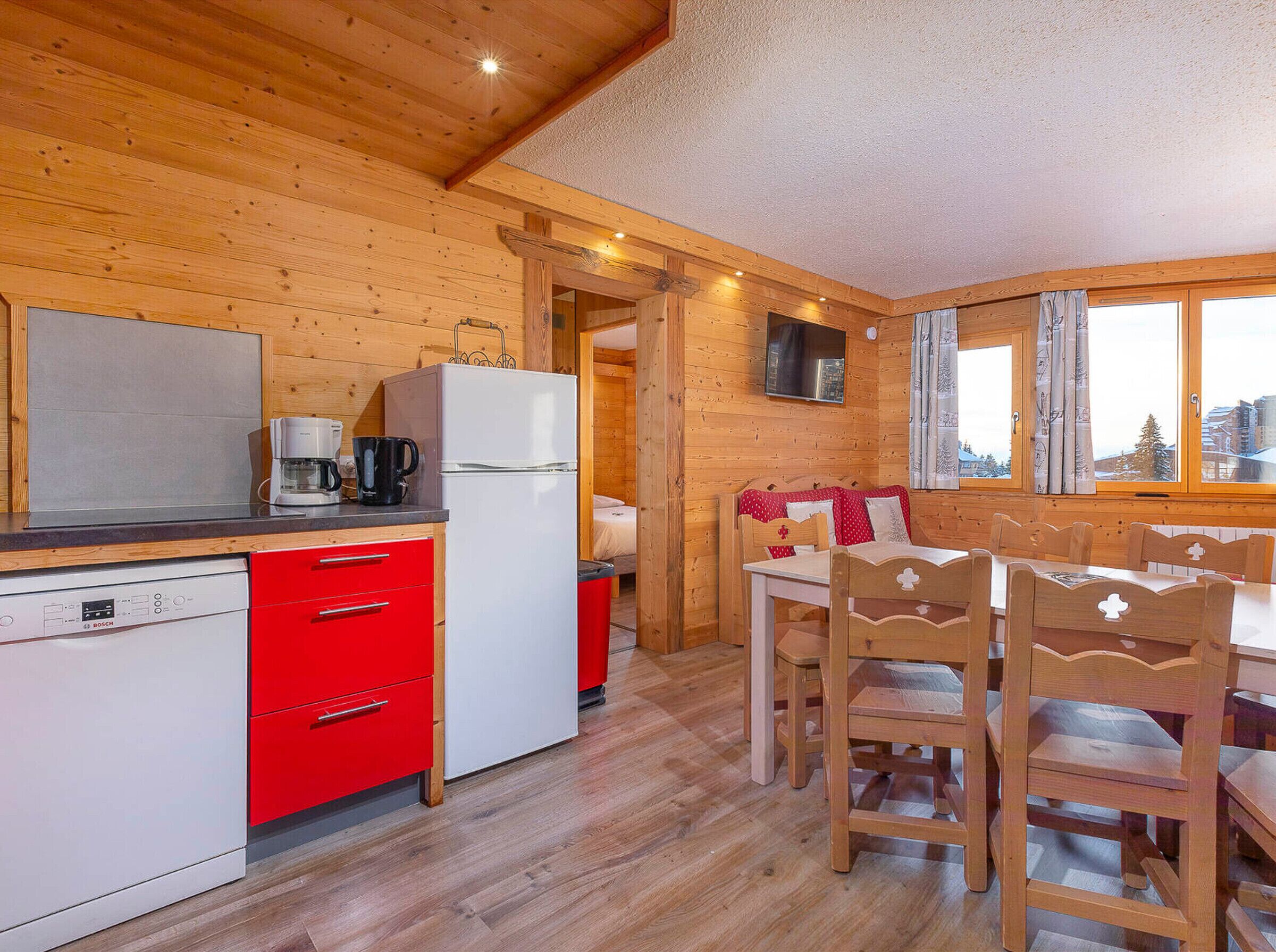 A cozy cabin-style room with wooden walls and a wooden ceiling. It includes a kitchen with a Bosch dishwasher, a white refrigerator, and a coffee maker. There is a dining area with wooden chairs and a table, and a window with curtains.