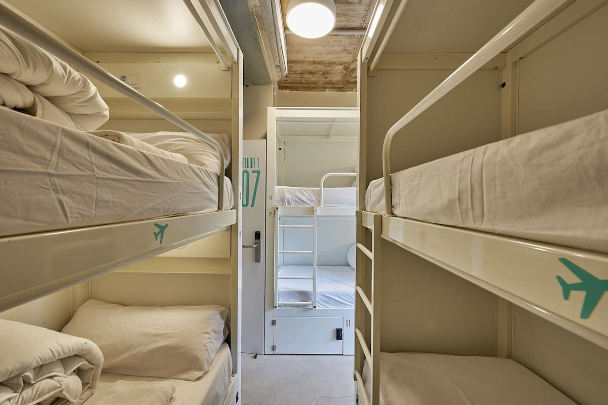 A narrow, enclosed sleeping area with bunk beds, a door leading to another room, and a ceiling light.
