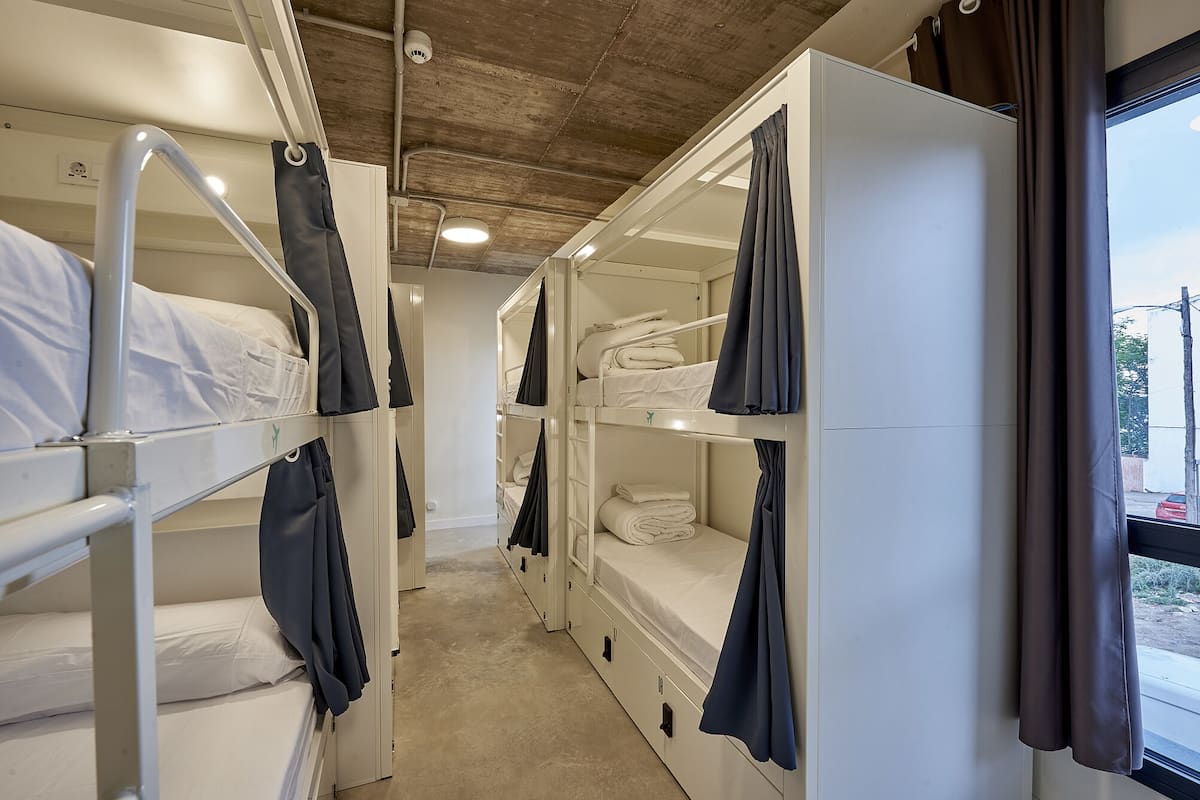 A narrow corridor with bunk beds, a window with curtains, and a concrete ceiling.