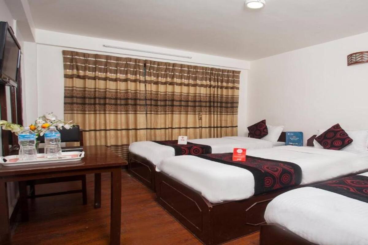A hotel room with two beds, a desk with a tray of water bottles, and a window with patterned curtains.