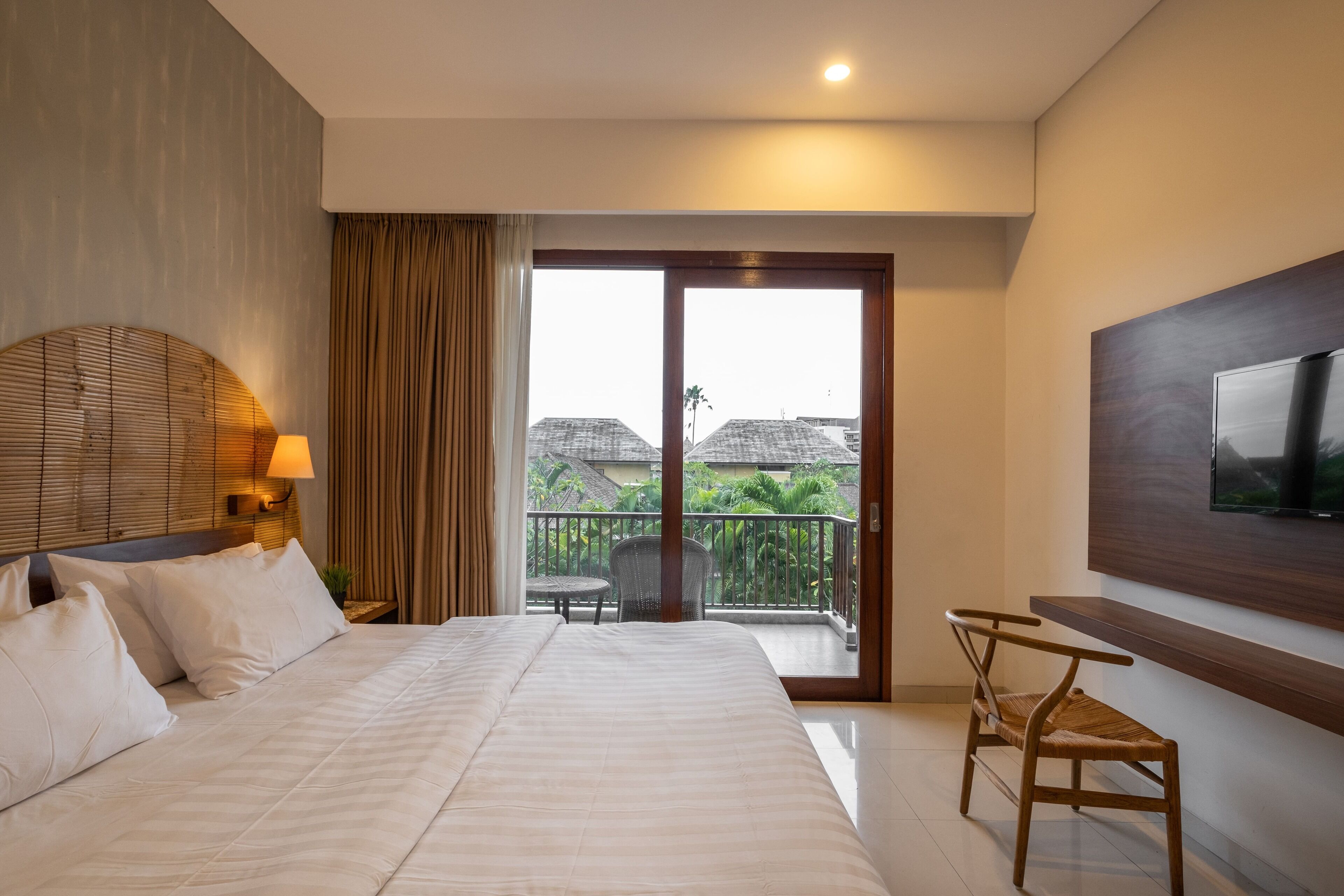 Habitación de hotel con una cama grande, un televisor montado en la pared, un balcón con una silla y vistas a edificios.