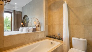 A modern bathroom with a bathtub, toilet, and a large mirror reflecting a bedroom with a bed and window. - Cove Tevana (Keroboken Kelod)