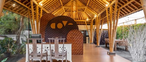 Un espacio al aire libre techado con techo de bambú, una amplia pared decorativa y una zona de comedor con mesa y sillas.