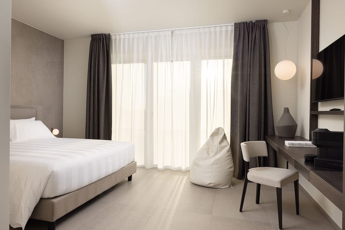 Moderni hotellihuone, jossa on sänky, työpöytä tuolilla, papupussi ja suuri ikkuna, jossa on verhot.