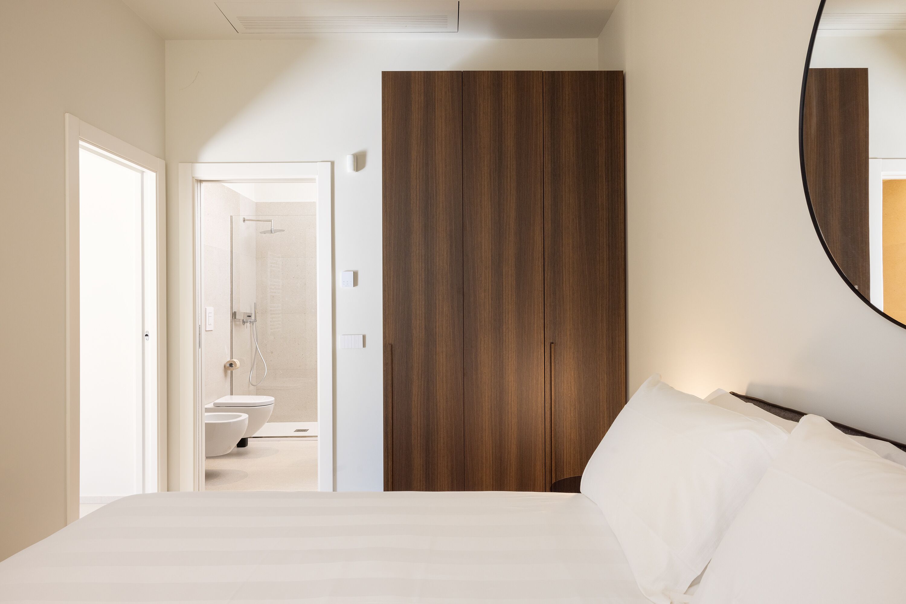 A bedroom with a bed, a wooden wardrobe, and a bathroom visible through an open door.