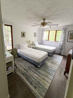 Une chambre avec deux lits, un ventilateur de plafond, un bureau avec une chaise et une fenêtre avec des rideaux.