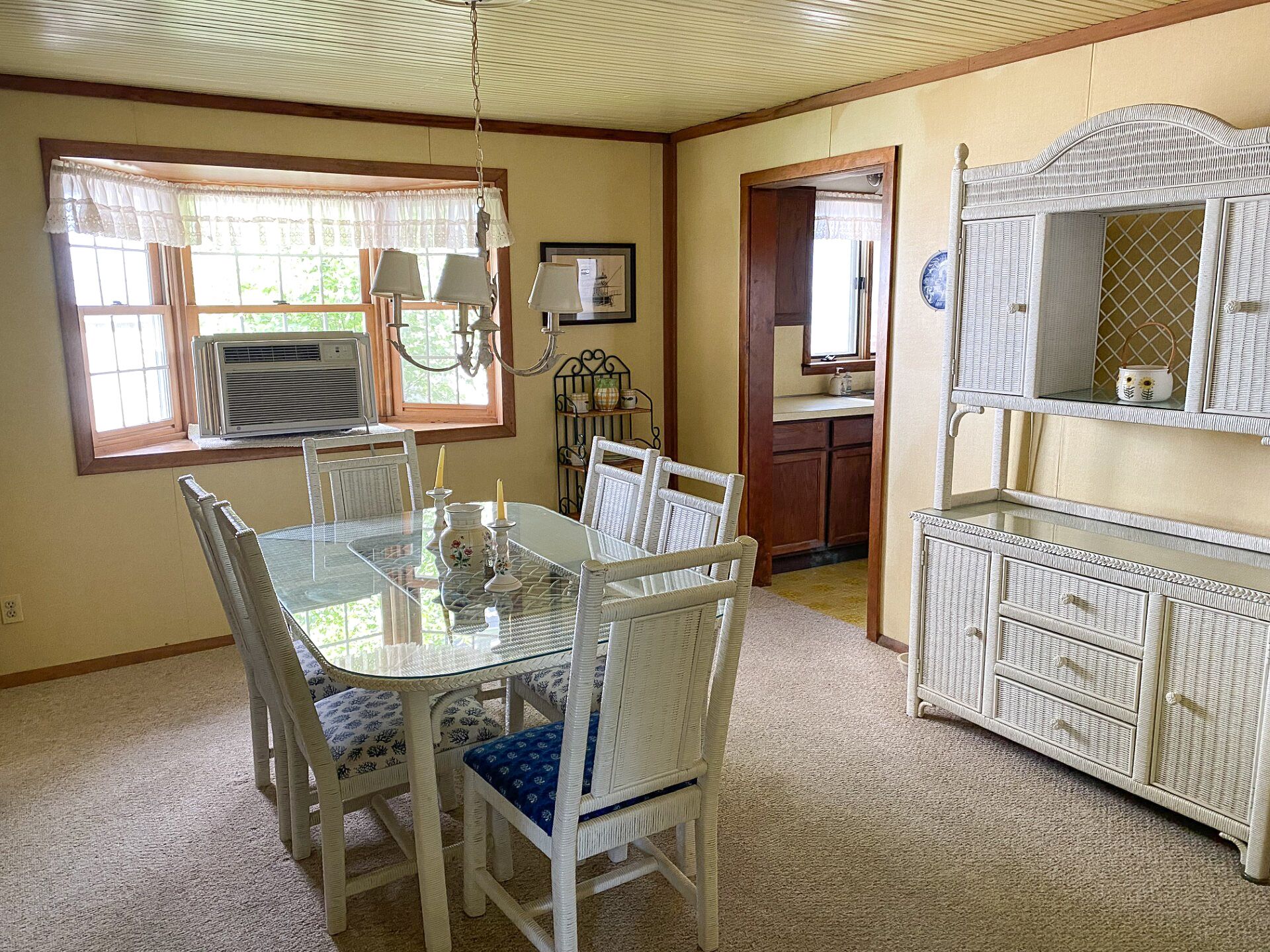 A dining area with a glass table and white wicker chairs, a kitchen with white wicker furniture, and a window with a white curtain.