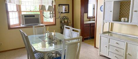 A dining area with a glass table and white wicker chairs, a kitchen with white wicker furniture, and a window with a white curtain.