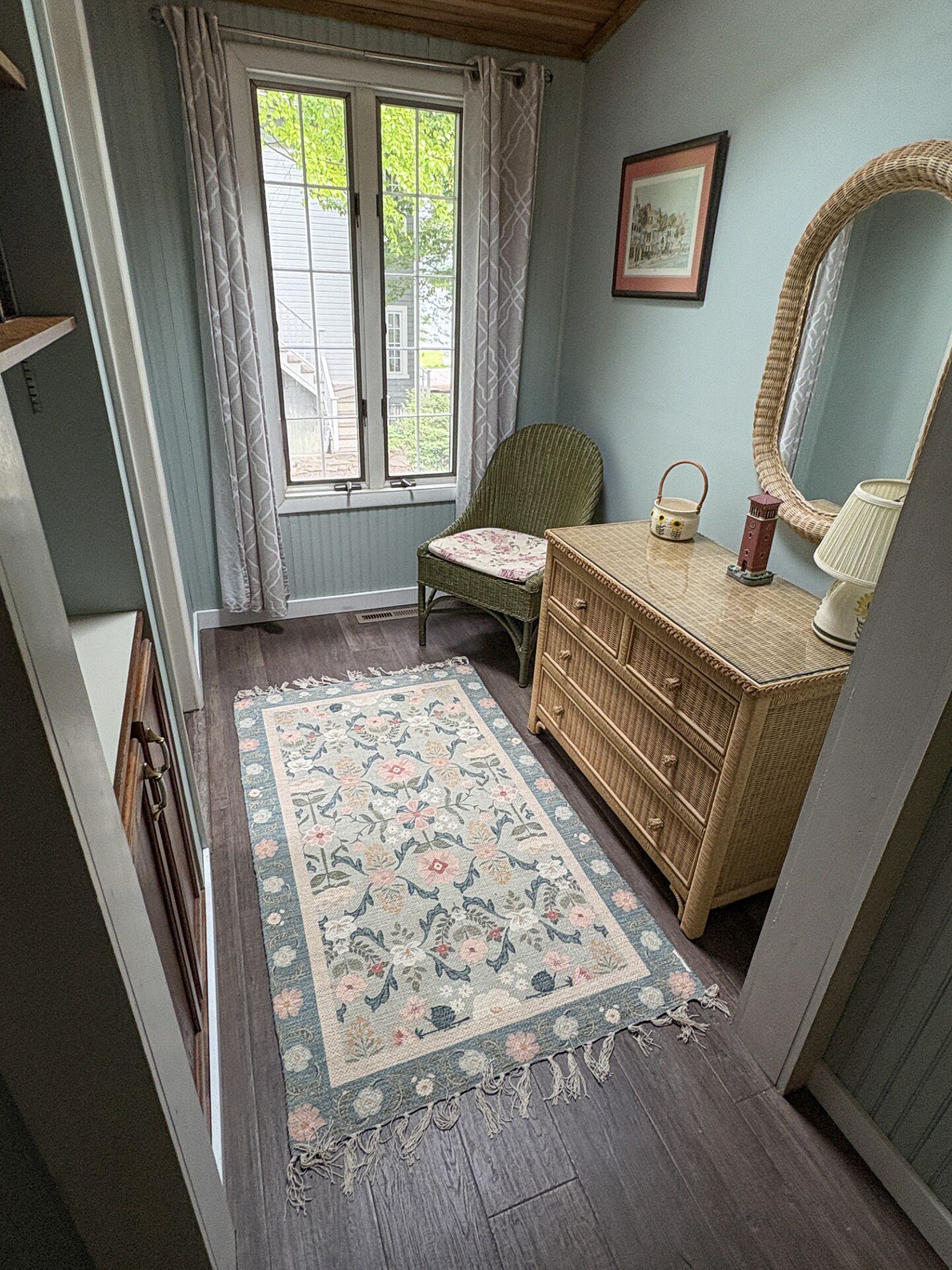 A room with a wooden dresser, a wicker chair, a floral rug, a mirror, and a window with curtains.