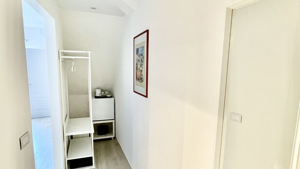 Um corredor estreito com uma porta branca à direita, um guarda-roupa branco com prateleiras e um quadro pendurado na parede.