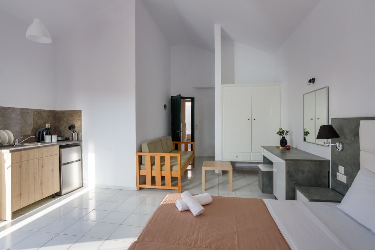 Una habitación de hotel moderna con área de cocina, una cama con cubierta marrón, una silla de madera, una mesita y un espejo.