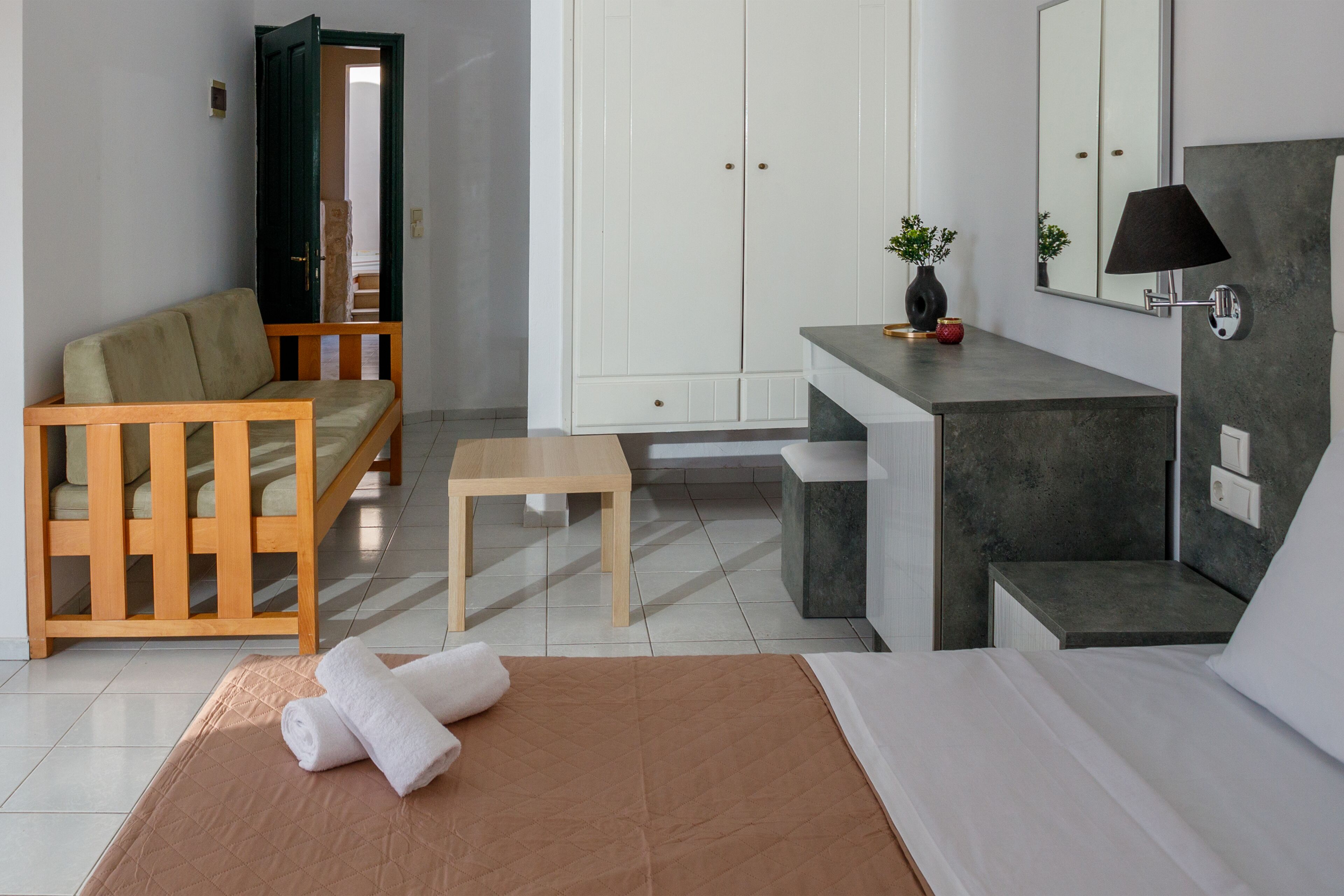 A modern hotel room with a bed, a wooden sofa, a small table, a vanity with a mirror, and a wardrobe.