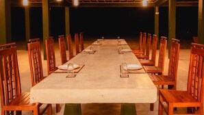 A long dining table with chairs, set for a meal, in a covered outdoor area with wooden beams and hanging lights.