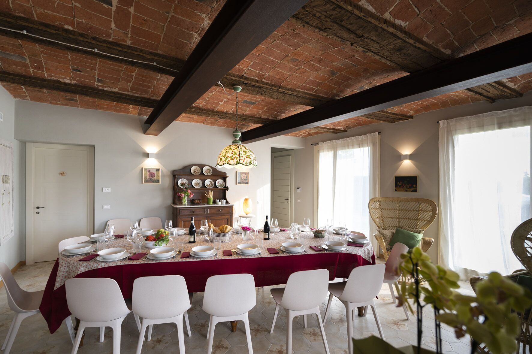 A dining area with a brick ceiling, white chairs, a table set for six, and a wicker chair by the window.