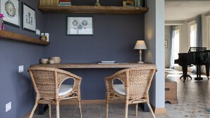 Una habitación con una pared azul oscuro, una estantería de madera con libros y objetos decorativos, dos sillas de mimbre, una mesa con una lámpara y un piano de cola al fondo.