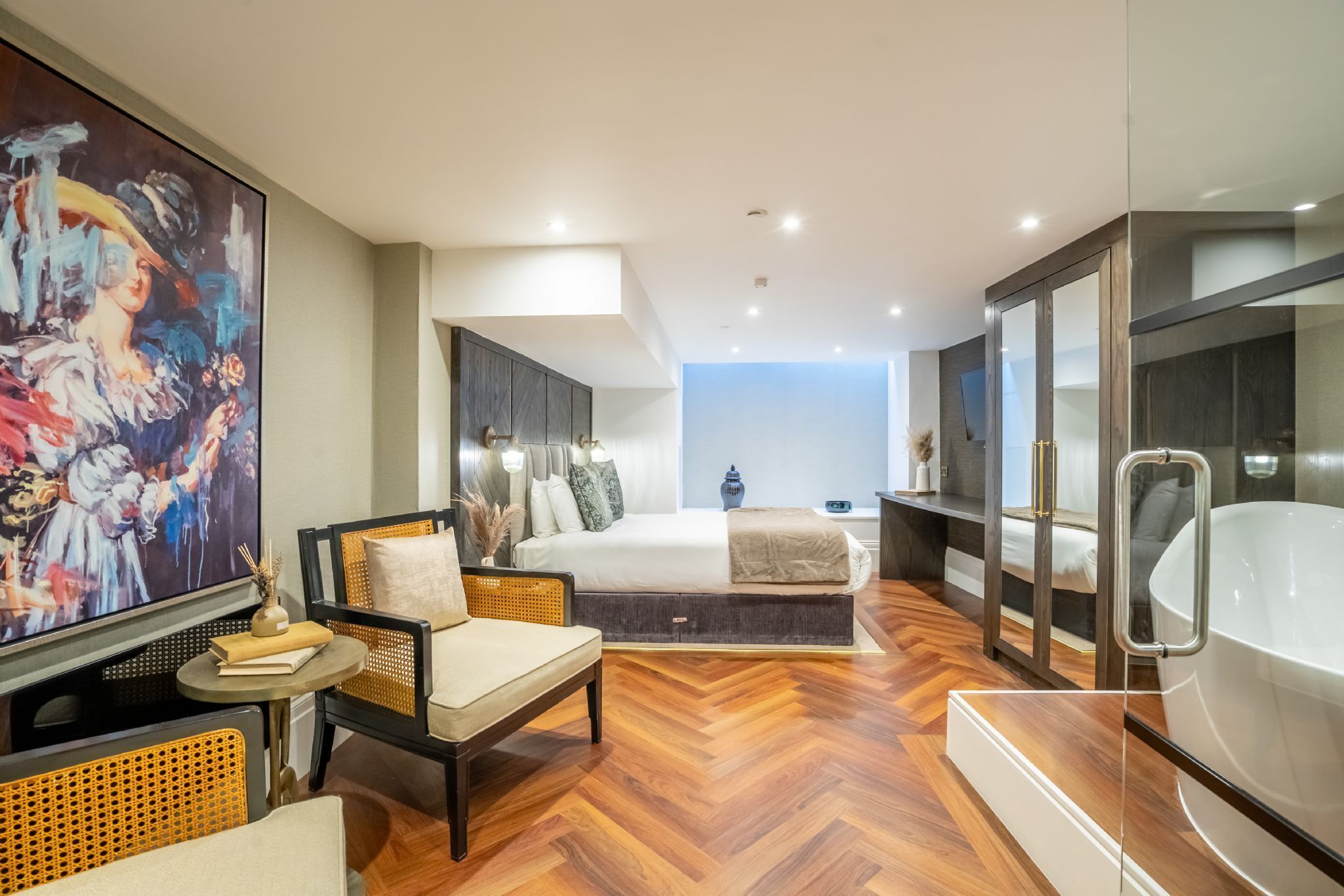 A modern hotel room with a large bed, a sitting area with a chair and table, a large painting on the wall, a bathtub, and a television.