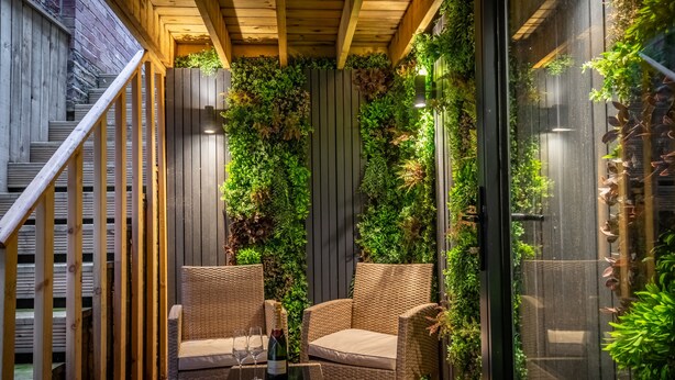A cozy outdoor seating area with wicker chairs and a glass table, surrounded by lush vertical gardens and wooden beams.