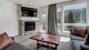 A modern living room with a stone fireplace, a large window with a view of a forest, and a grey sofa with brown cushions. - Clearwater Lofts Luxury 1 Bed Condo Near Lakeside Village (Keystone)