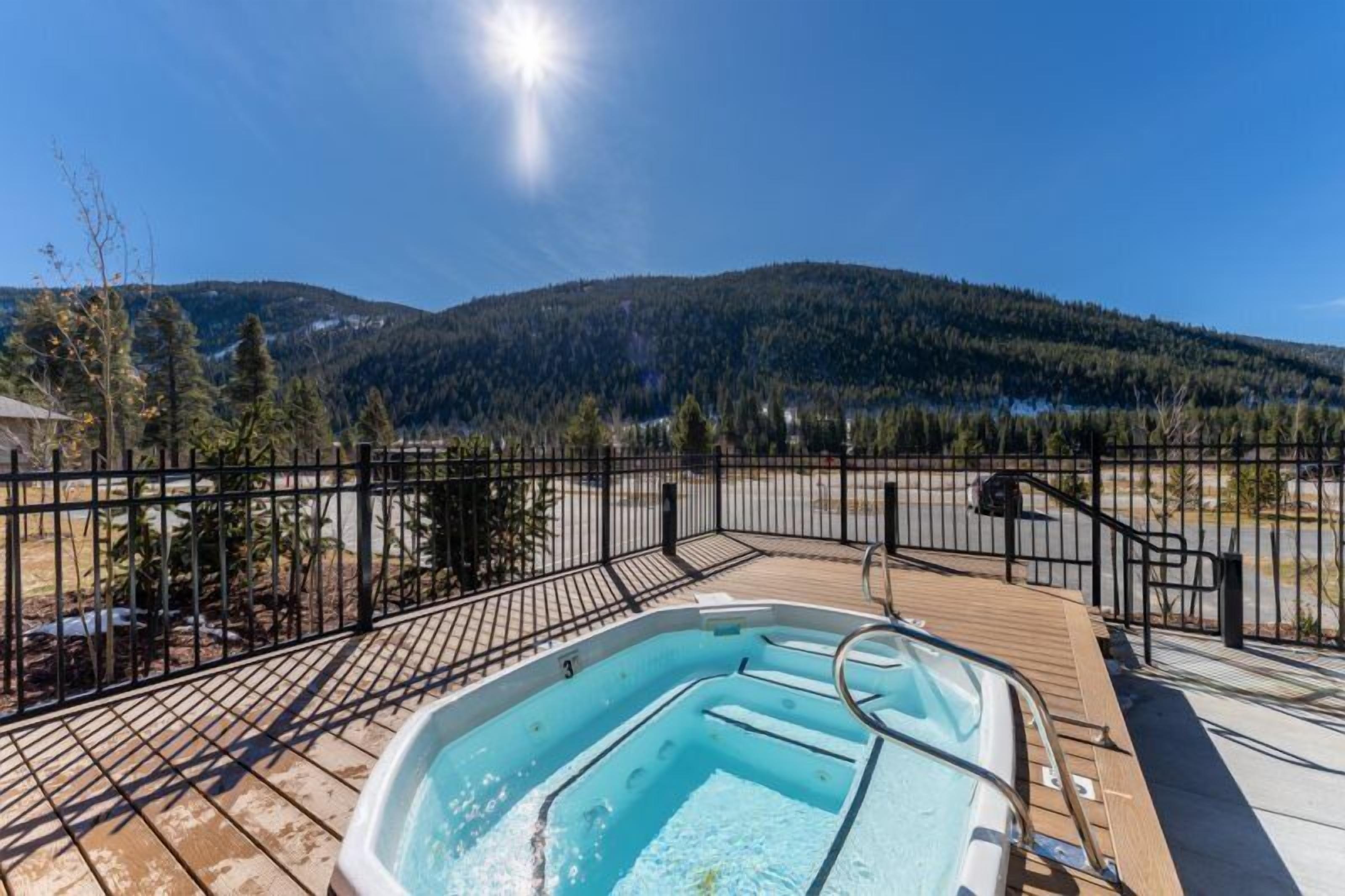 A hot tub on a wooden deck with a metal fence, surrounded by trees and a mountain in the background.