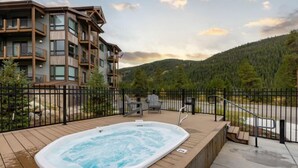 A hot tub on a wooden deck with a view of a multi-story building and a forested hillside. - Clearwater Lofts Luxury 1 Bed Condo Near Lakeside Village (Keystone)