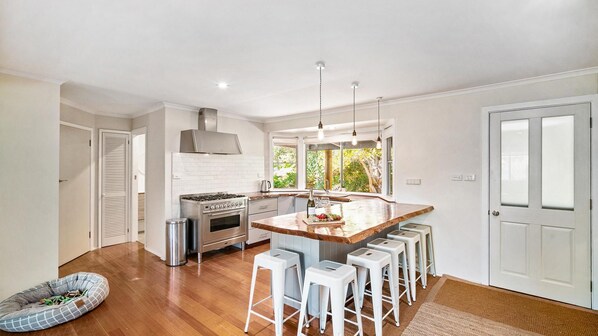 A modern kitchen with a central island, bar stools, a dog bed, and a large window providing a view of the outdoors.