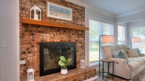A living room with a brick fireplace, a beige sofa, and a wooden floor. - Discover Sea Glass: a cozy Oak Island retreat with 3 bedrooms and pet-friendly amenities! (Oak Island)