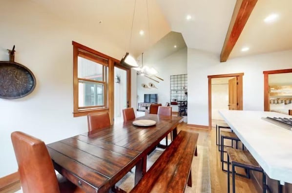 A dining area with a wooden table and benches, a kitchen in the background, and a large window.