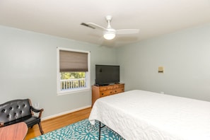A bedroom with a bed, a television on a dresser, a ceiling fan, a window with blinds, and a chair.